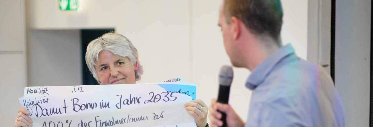 Eine Bürgerin hält ein großes Flipchart-Plakat hoch mit Forderungen für die Mobilitätswende, ein Mann mit Mikrophon liest es vor