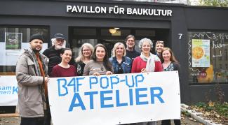 10 Personen stehen vor dem Pavillon. Sie lächeln und halten ein Banner hoch mit derm Text: B4F-Pop-Up-Atelier